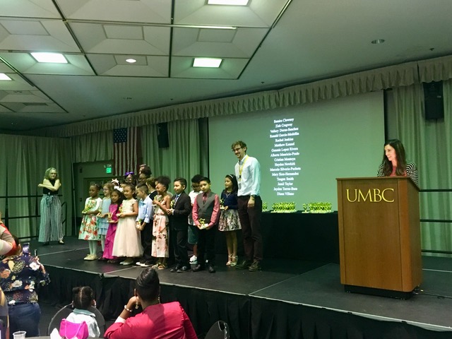 A UMBC male graduate student stands on stage with a mixed group of local elementary school age students for a celebration.