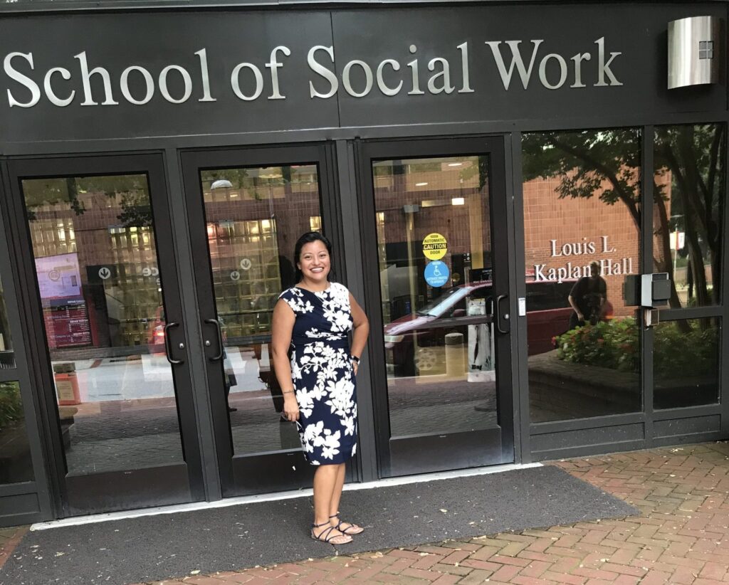 Woman in black and white floral dress and sandals stands outside a building reading 