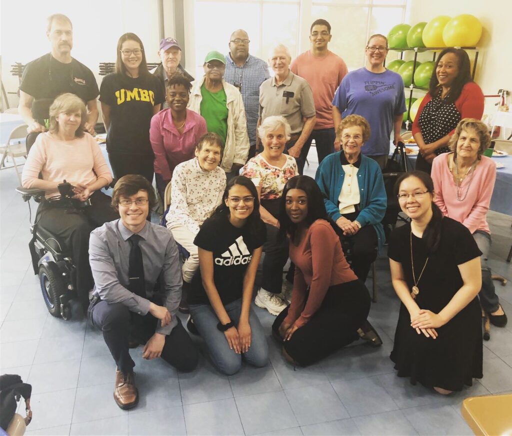 A group of nineteen people, some standing some sitting, inside a community gym with a rack of therapy balls in the background.