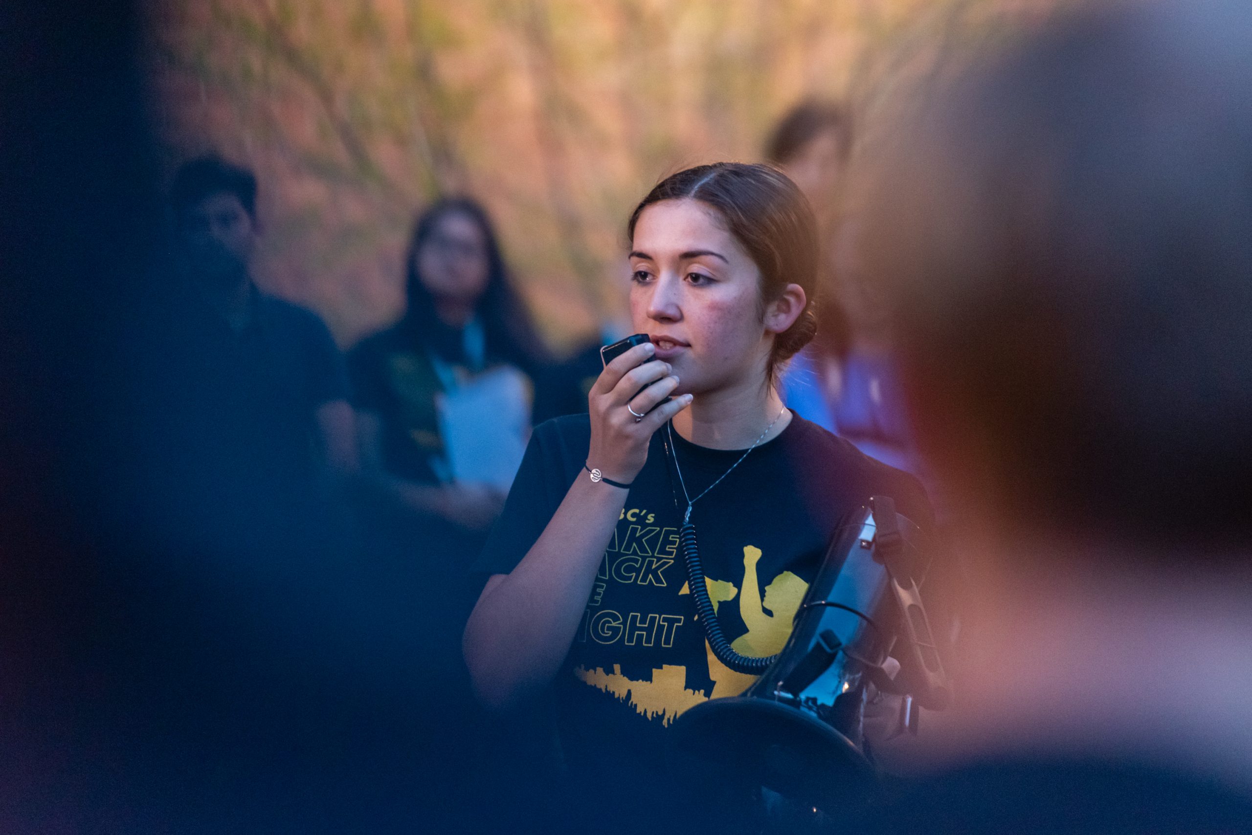 Woman wearing "Take Back the Night" t-shirt speaks into a microphone at the center of a group.