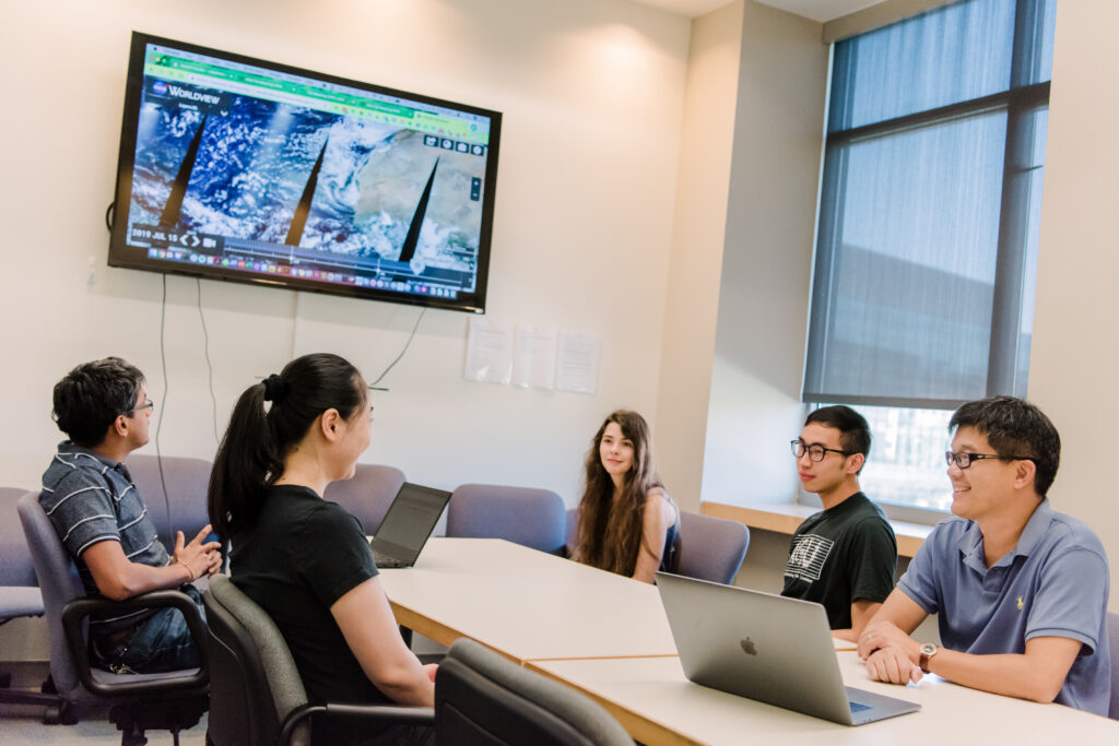 Group of five people sits at a conference table. Large screen on the wall shows satellite imagery of Earth. 