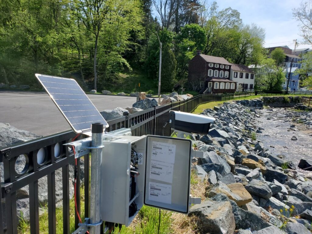 Box of electronic equipment with a solar power panel. Outdoor photo with road, plants, and homes in the background.