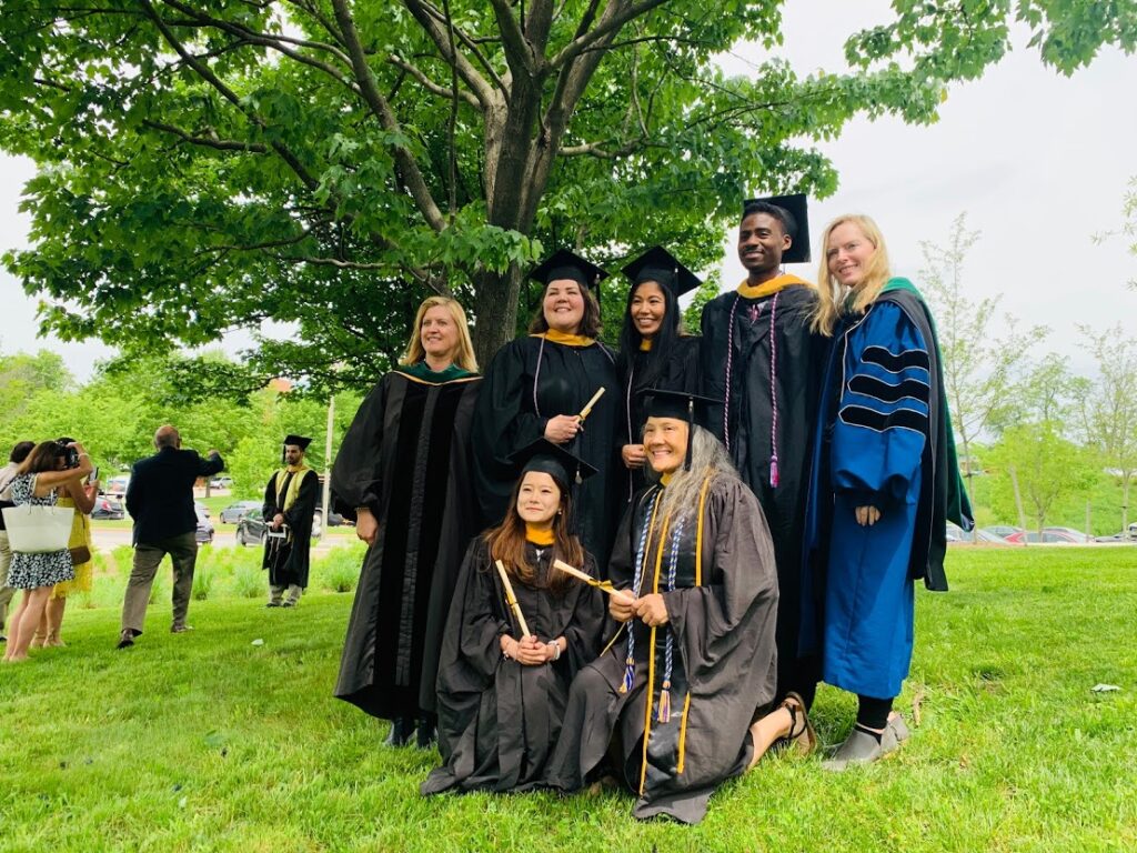 A group of five graduate students and two professors wearing caps and gowns standing under a tall tree in springtime.