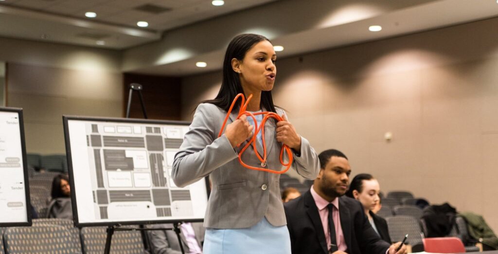 Young woman with long dark hair wearing a grey blazer and light blue skirt, standing and holding twisted orange wire,
