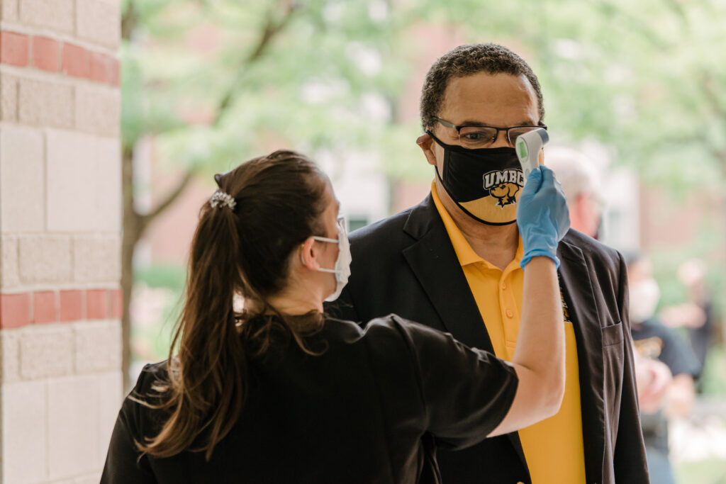 Man in polo shirt and blazer, wearing UMBC face mask, gets temperature taken by medical professional outdoors