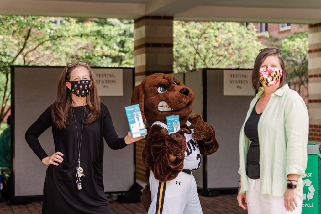 Two women stand on either side of a person dressed in a dog mascot costume. The women wear face masks. One holds up a thermometer in a package.