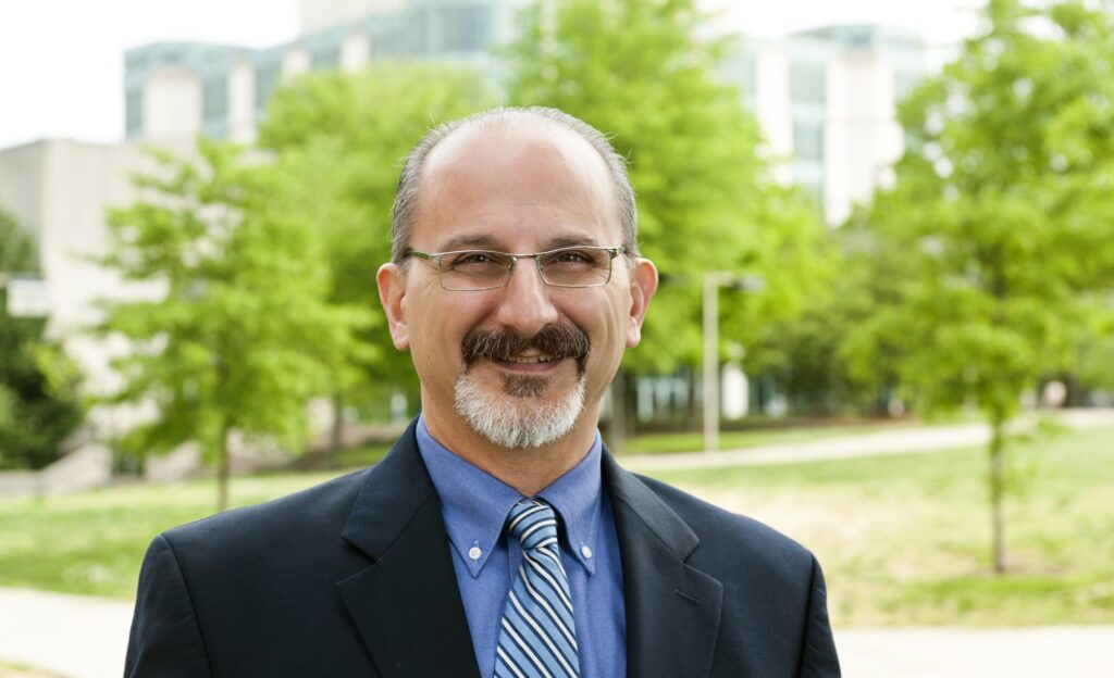 Man with glasses, mustache and short beard is wearing a blue suit and striped tie smiles at camera with green trees in the foreground.