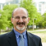 Man with glasses, mustache and short beard is wearing a blue suit and striped tie smiles at camera with green trees in the foreground.