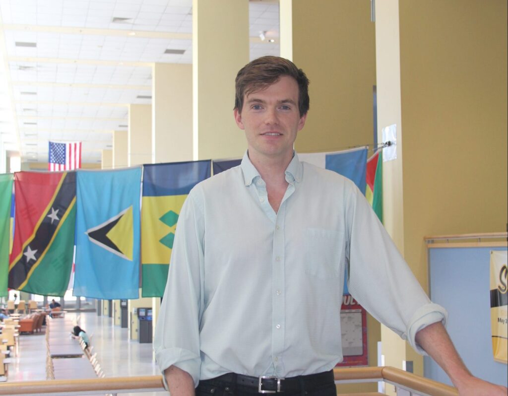 Man with brown hair, wearing a white dress shirt and black pants, with a row of international flags hanging in the background, smiles at camera.