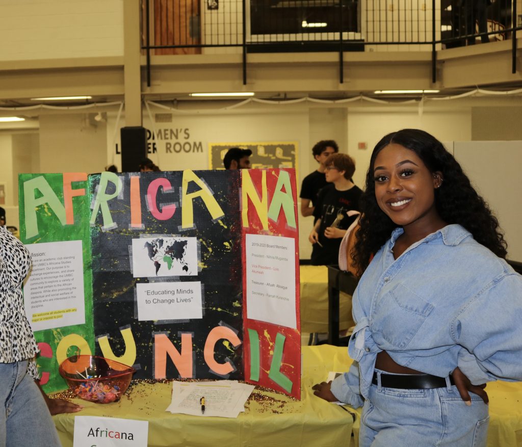 A female college student wearing jeans and a denim log sleeve shirt smiles while standing in front of a poster board that says Africana Council.