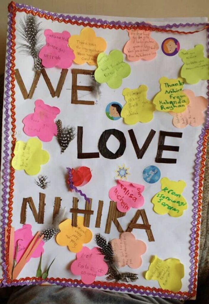 A white paper made into a card is decorated with yellow, pink, and peach bear cut outs with names written inside the cut-outs, purple and orange zigzag strips are pasted on the right and left sides of the paper, with the words We Love Niihira cut out of different colored paper and  pasted in the middle of the card.