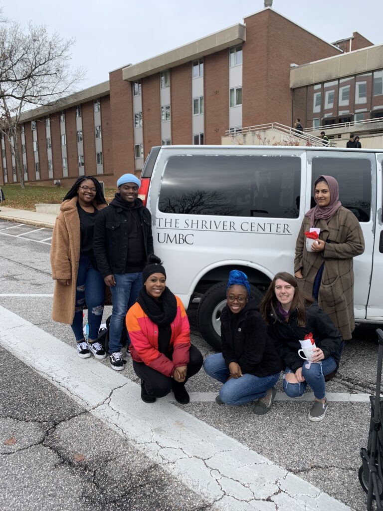 A group of six college students wearing winter jackets, stand in front of a a white van that has lettering that reads The Shriver Center UMBC. Three of them are kneeling on the pavement.