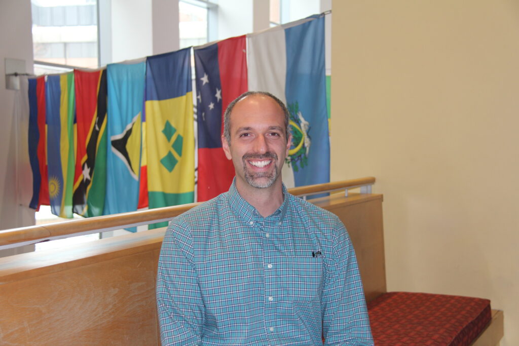 Man with grey hair and goatee, wearing a white and blue checkered shirt, stands smiling at camera with international flags in the background.