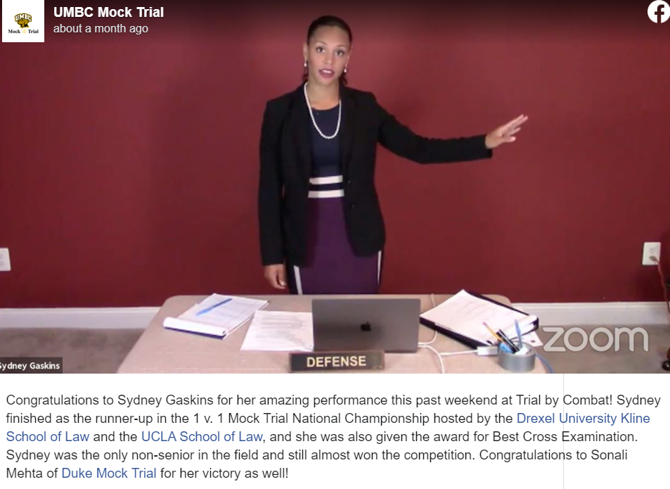 A UMBC Mock Trial Facebook post of a young woman with black hair pulled back wearing a dark purple dress with white stripes on the waist, a black blazer, and pearl  necklace point to her left and stands behind a beige table that has a laptop and papers spread across it. 
