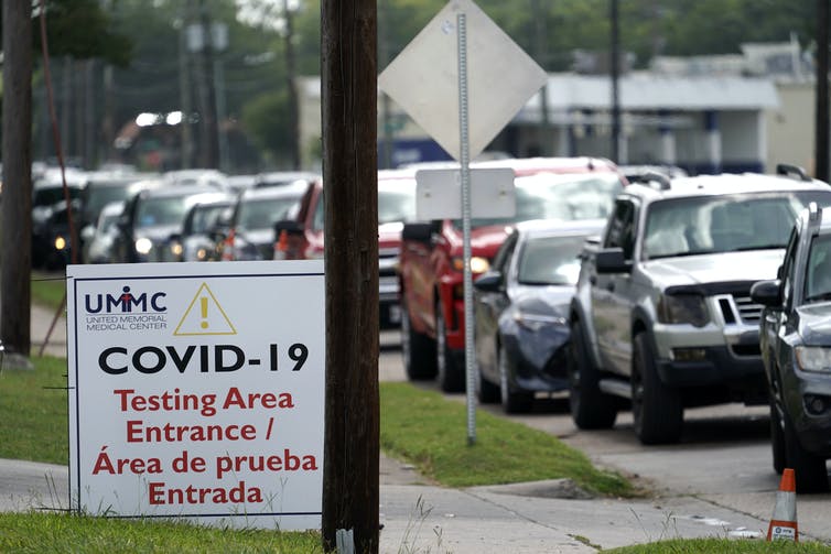 A line of people waiting in cars in front of a sign for COVID-19 testing.