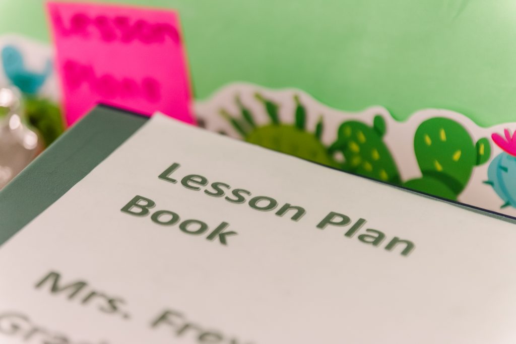 A green binder laying flat with a white paper on top that has the words "lesson plan book" written in green. There is green paper with a border of cacti in the background.