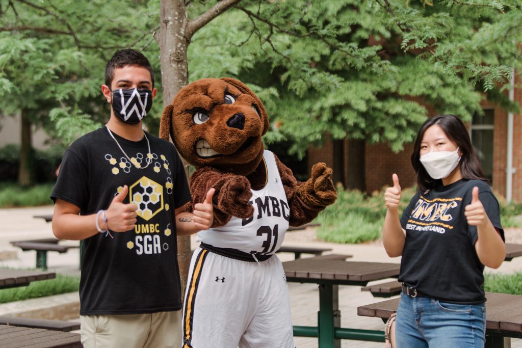 A man and a woman with face masks on next to a dog mascot.