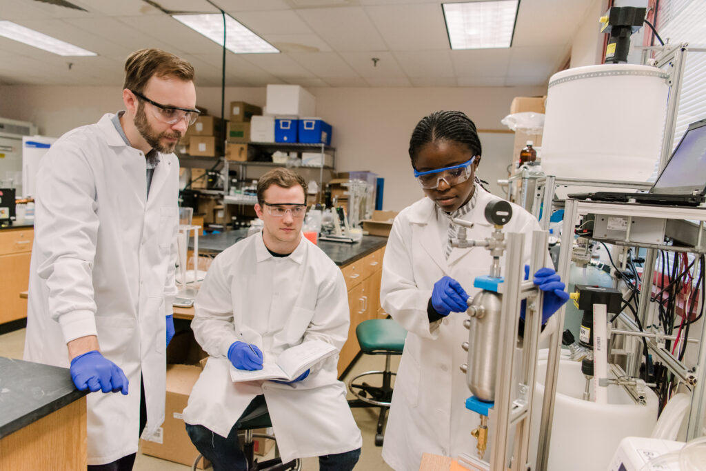 Middle-aged white man stands with a young white man and young Black woman in a lab. They all wear protective goggles and lab coats. The young woman demonstrates research equipment.