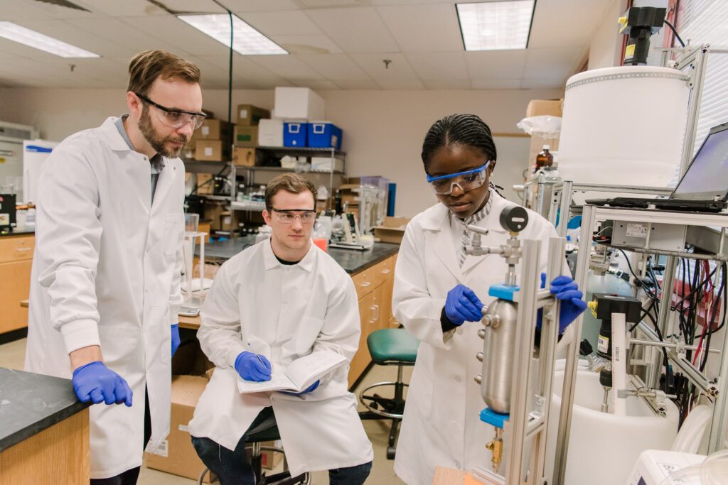 A student uses research equipment in an environmental engineering lab. Another student takes notes while a professor observes.