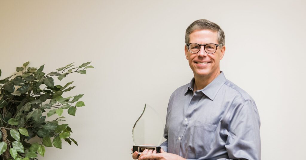 A man with grey hair and a grey mustache wearing glasses and a blue and white checkered dress shirt smiles at the camera. There is a wall behind him with the word truth written on it in white.