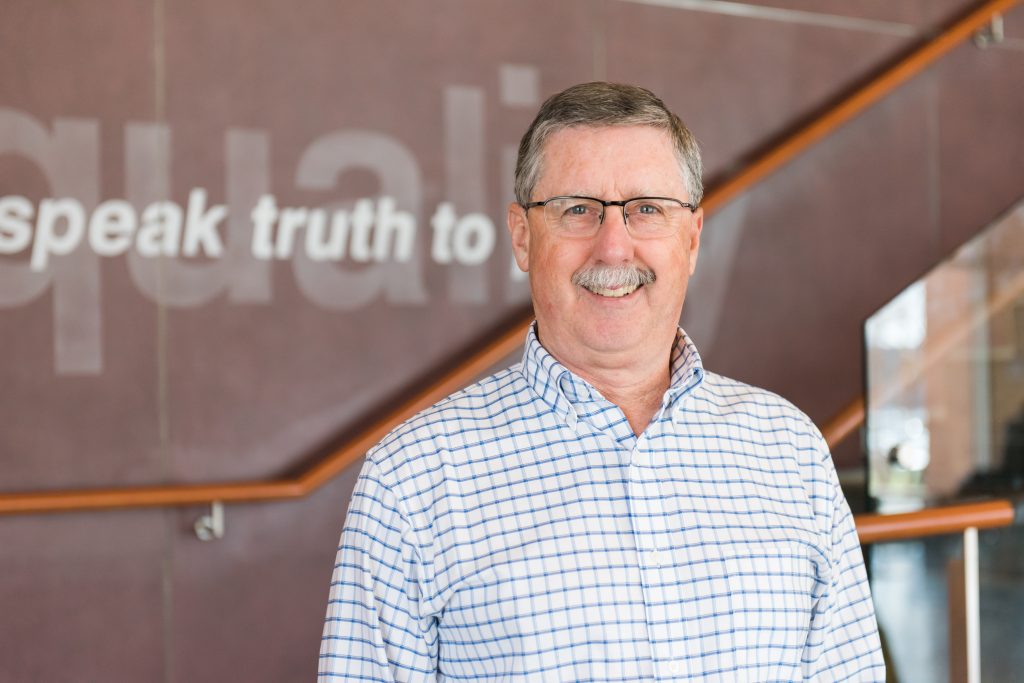 A man with grey hair and a grey mustache wearing glasses and a blue and white checkered dress shirt smiles at the camera. There is a wall behind him with the word speak truth to written on it in white.