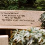 A picture of beige stone bench in the sunlight with a quote by Walter Sondheim etched into the back of the bench, "Try to understand someone else's position and not assume you know what's best for others."