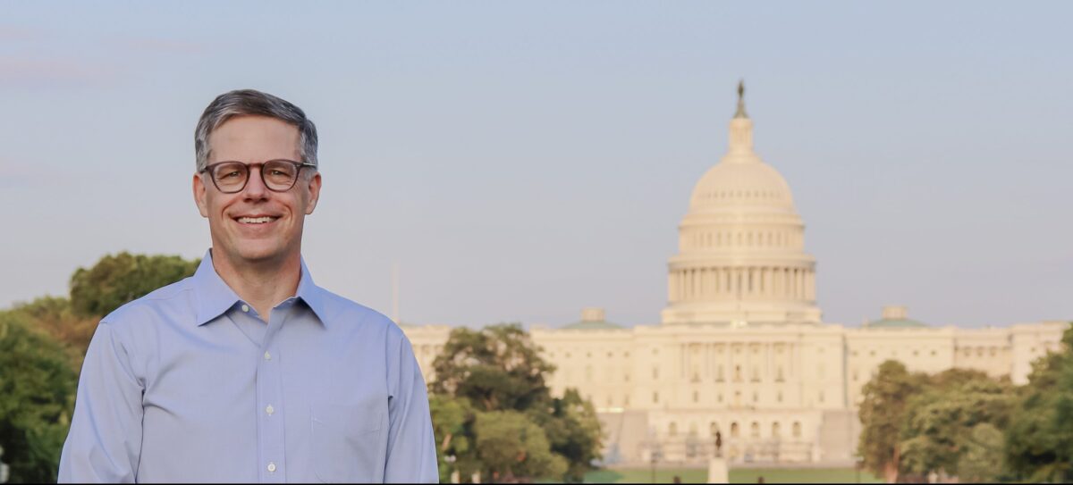 Man with greying short hair, wearing glasses and a blue dress shirt, smiles at the camera with the U.S. Capitol building in the background.