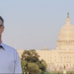 Man with greying short hair, wearing glasses and a blue dress shirt, smiles at the camera with the U.S. Capitol building in the background.