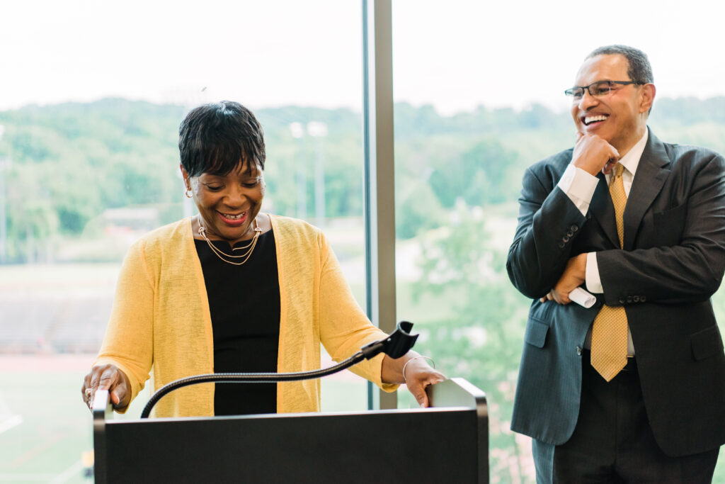 Woman stands at podium, smiling. Man stands to the side, behind her, in a suit, also with a friendly smile.