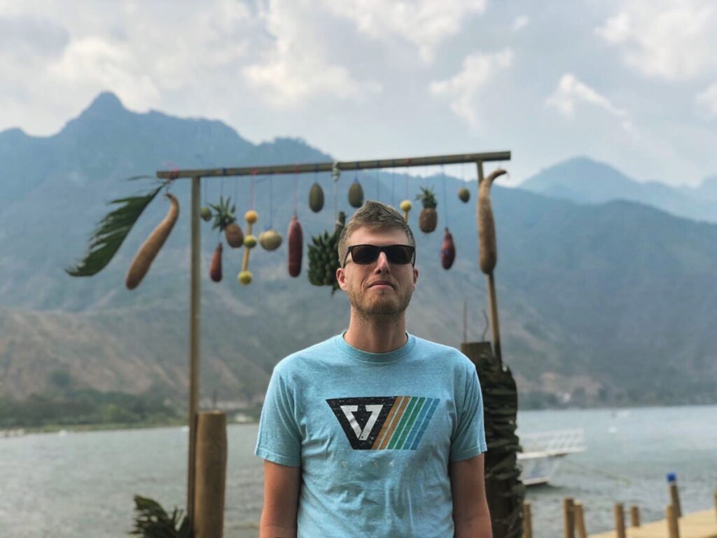 Young man with short brown hair, wearing black sunglasses and a light blue shirt with a multicolored design, stands in front of some objects hanging from a wooden pole and with mountains and a body of water behind him. 