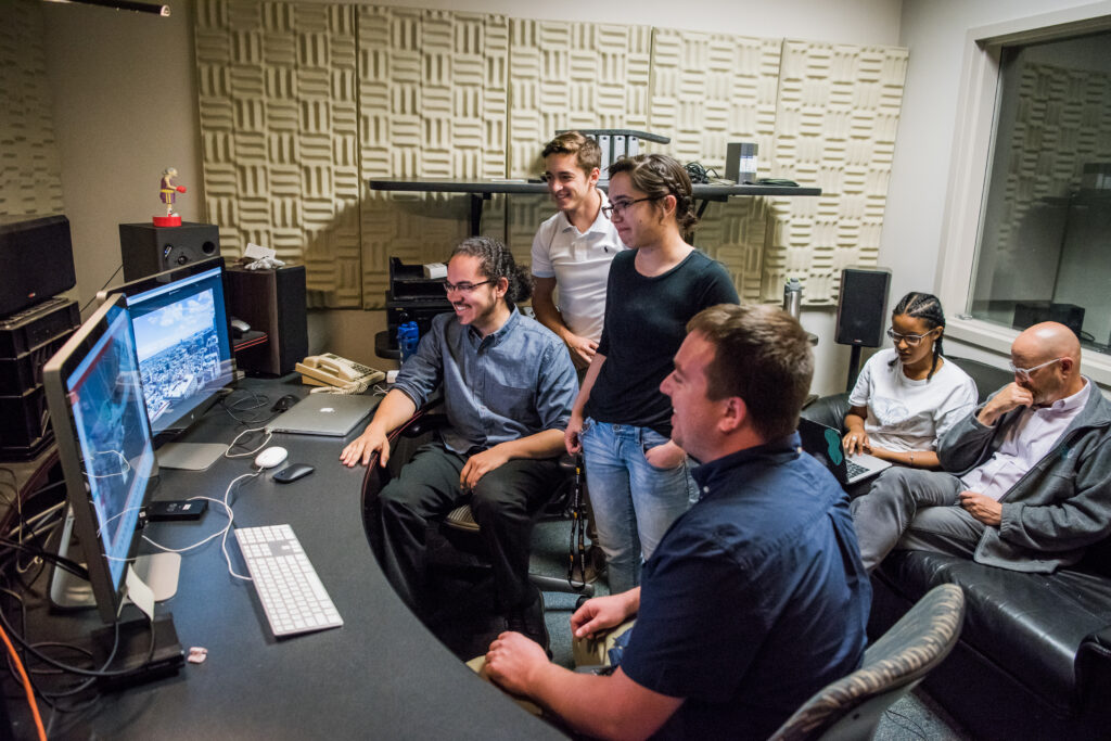 Four students gather around monitors, with a professor and student sitting in the background.