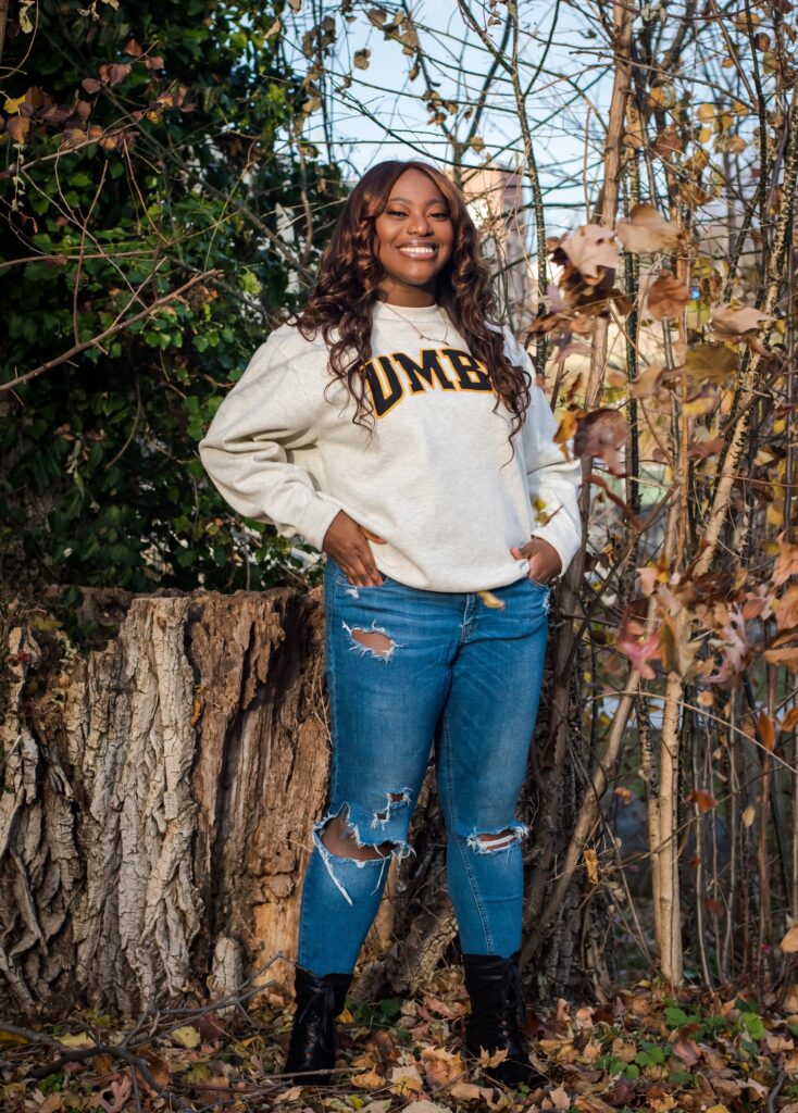 Young woman wearing ripped blue jeans, a white sweatshirt with the words UMBC written in the middle in black, and black boots, stands in front of some trees and shrubs while smiling at camera.