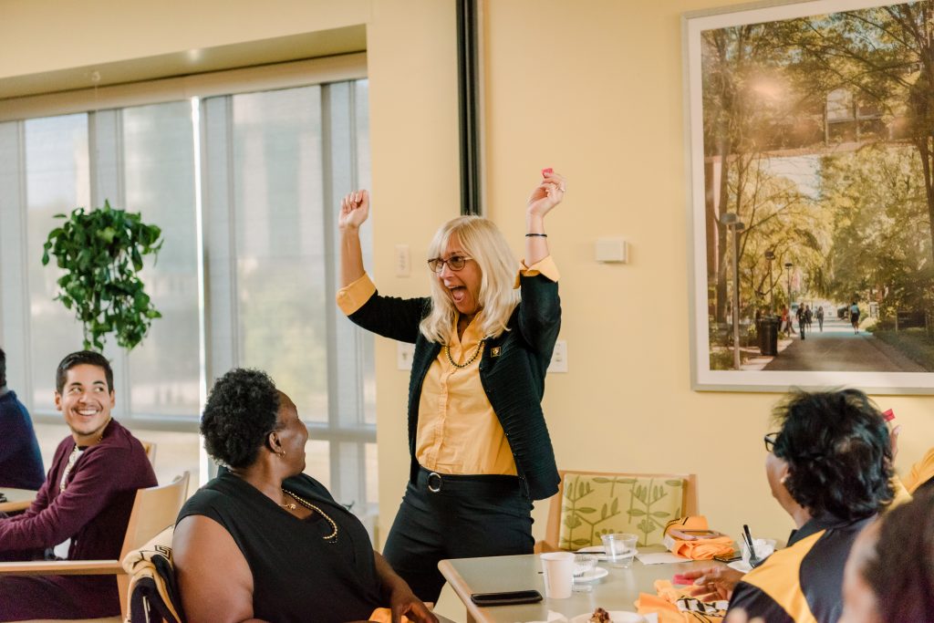 Blonde woman wearing black and gold with arms up celebrating, people sitting around her watch and smile.