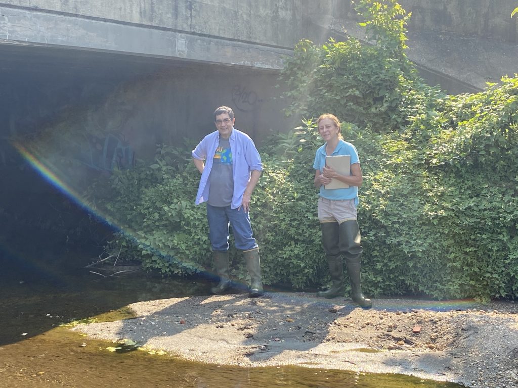 Two researchers stand below a concrete bridge, behind them is a giant green shrubbery and infront of them is sand and water.