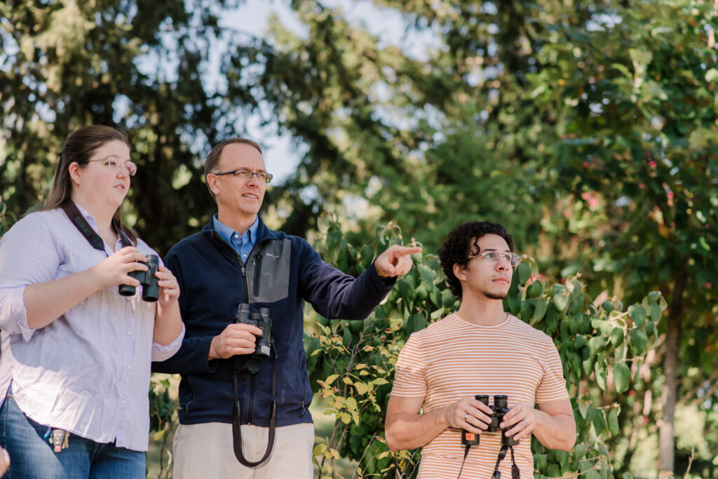 One woman and two men hold binoculars and stand in front of a field of trees.