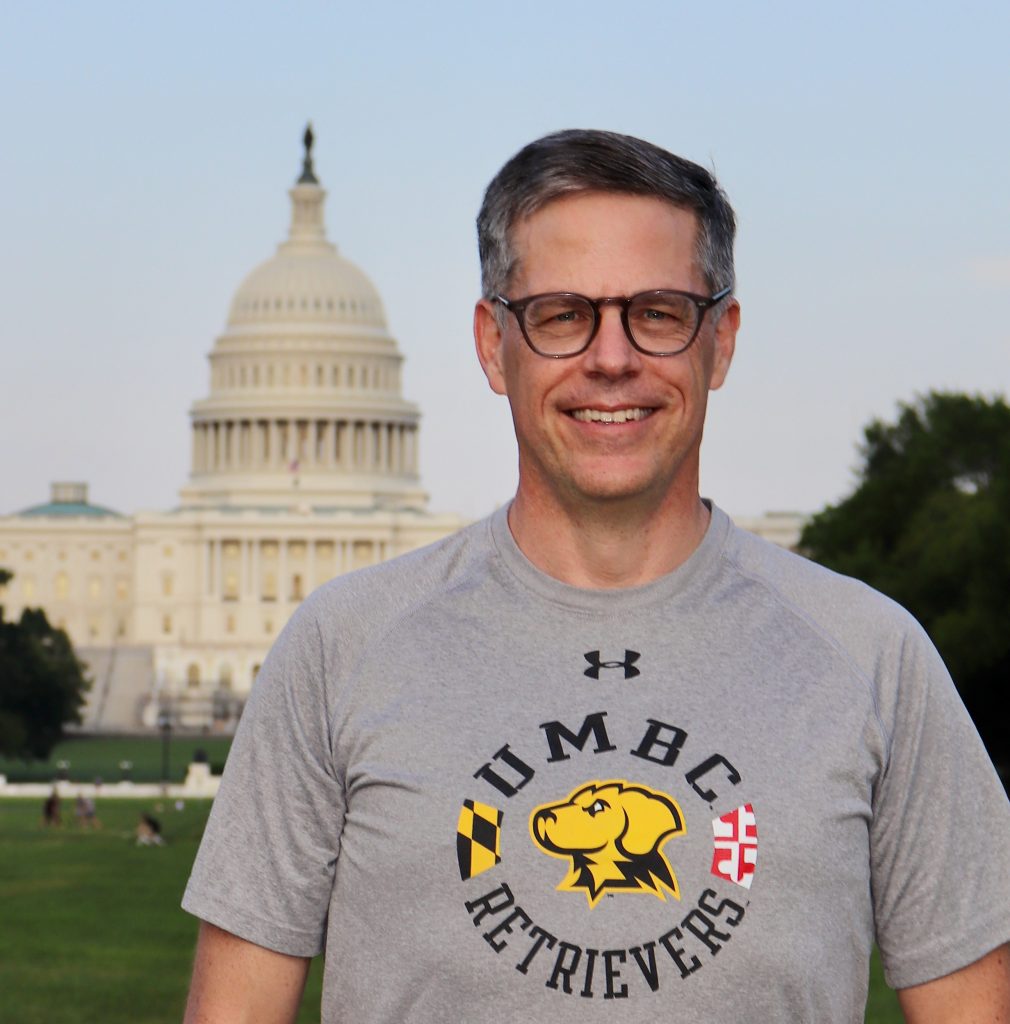 A man with greying hair wairing glasses and wearing a t-shirt that says UMBC Retrievers with a picture of a Chesapeake Bay Retriever with the U.S. Capitol in the background.