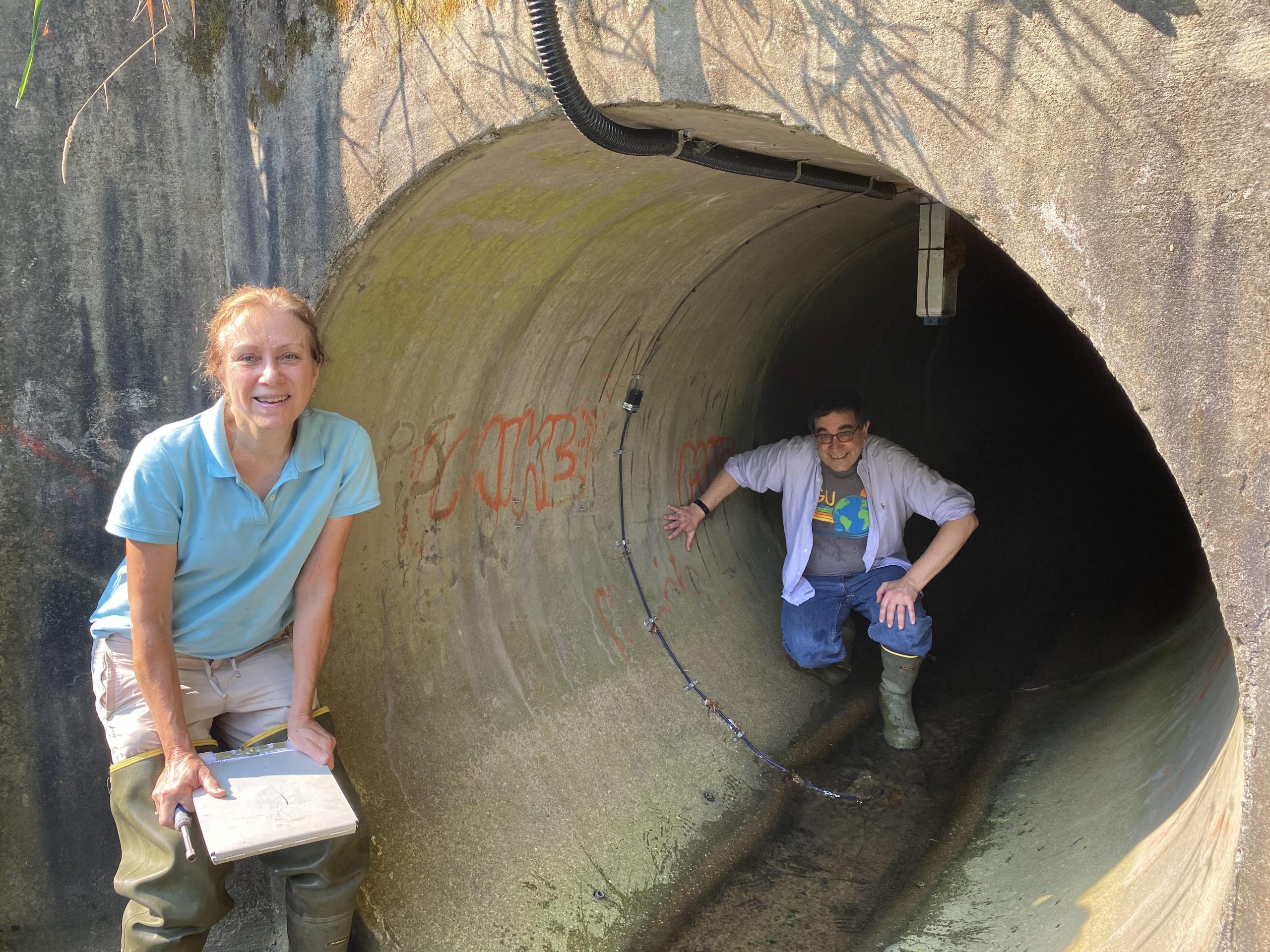 Man and woman in field research attire stand next to and inside a concrete tunnel at a research site.