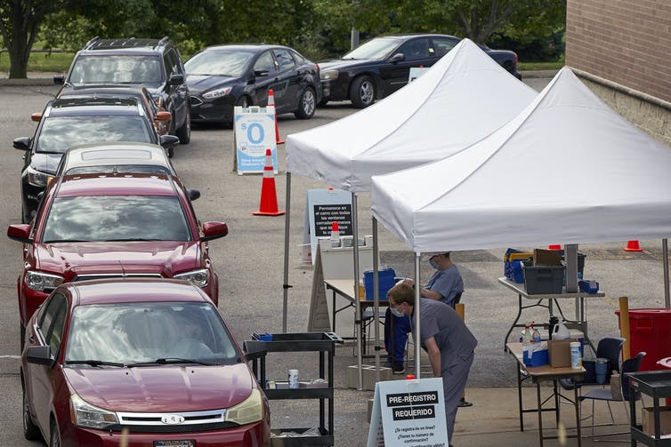 People waiting in a line of cars with health care workers standing under pop-up shade structures.