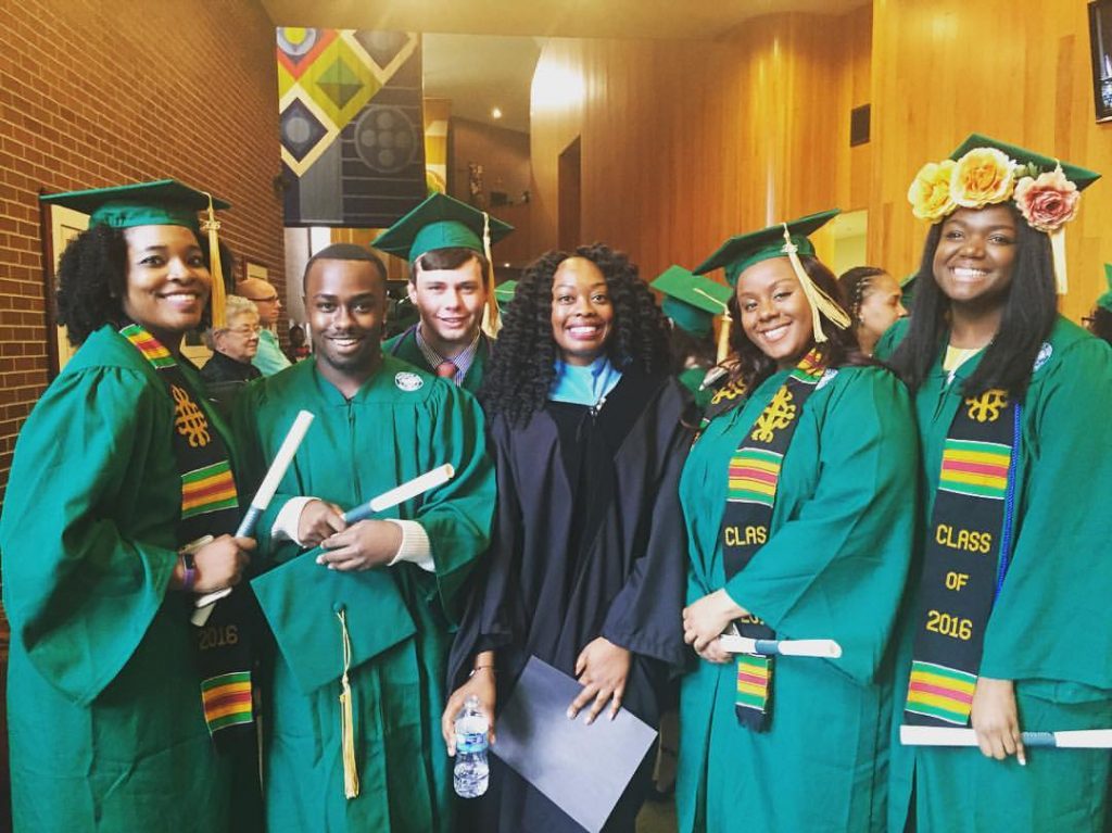 Six students wearing graduation caps and gowns, four in dark green with multicolored stoles reading.