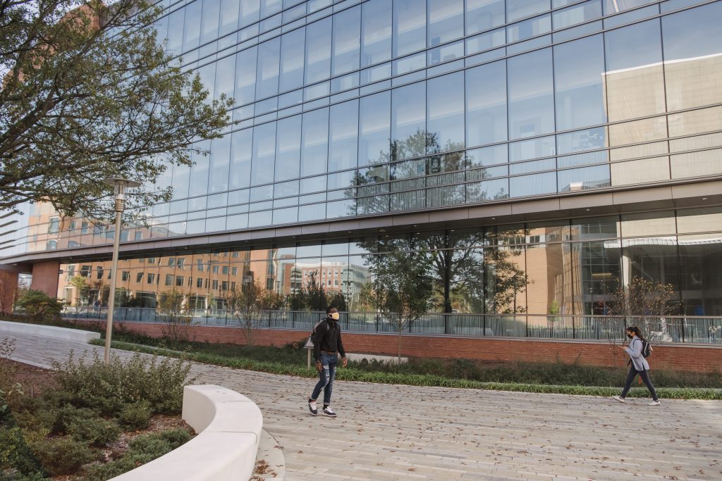 Two students wearing face masks walk in front of a large building with a reflective glass exterior, surrounded by fall foliage.