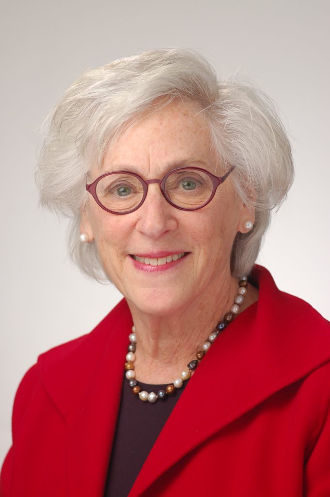 A White woman with grey hair, wearing red glasses, a red blazer with a black blouse, and a pearl necklace of different colors smiles at the camera.
