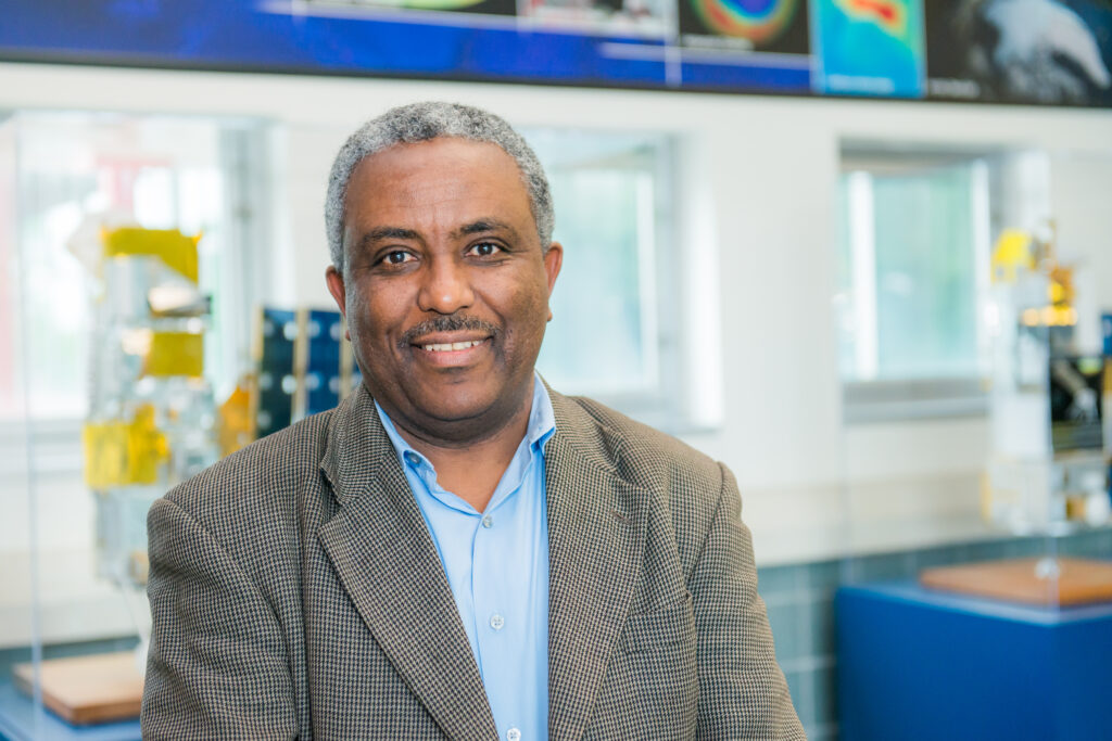 Middle-aged black man with graying har and a mustache smiles while posing in front of a display of space research equipment