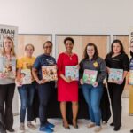 A group of woman of different races and ages stand next to eachother smiling at the camera while holding hard cover picture books in their hands. They are standing in between one white and one yellow standup banner with the words Sherman Center written on them.