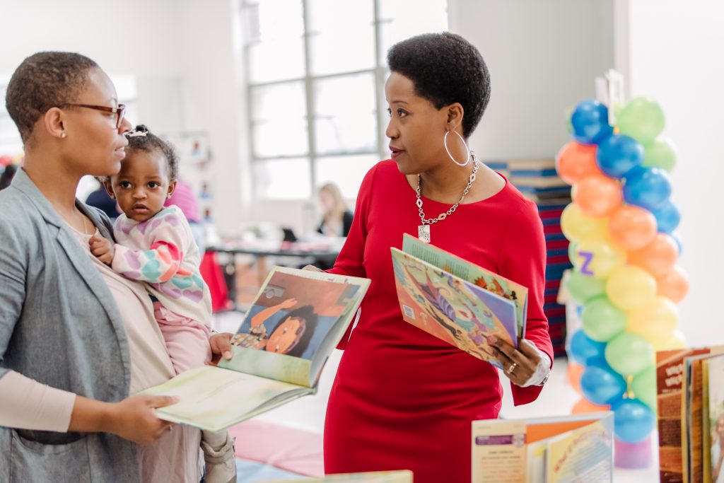 Two black women, one wearing a red dress and the other a beige shirt with a grey dress jacket, talk to each other while holding children's books. The woman with the grey jacket is holding a toddler. There is a column of balloons behind them.
