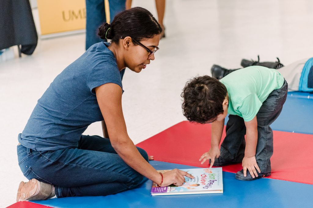A woman wearing jeans and a blue t-shirt kneels on a blue and red mat and points to a hard cover children's book on the mat. A toddler bends over to look at the book.