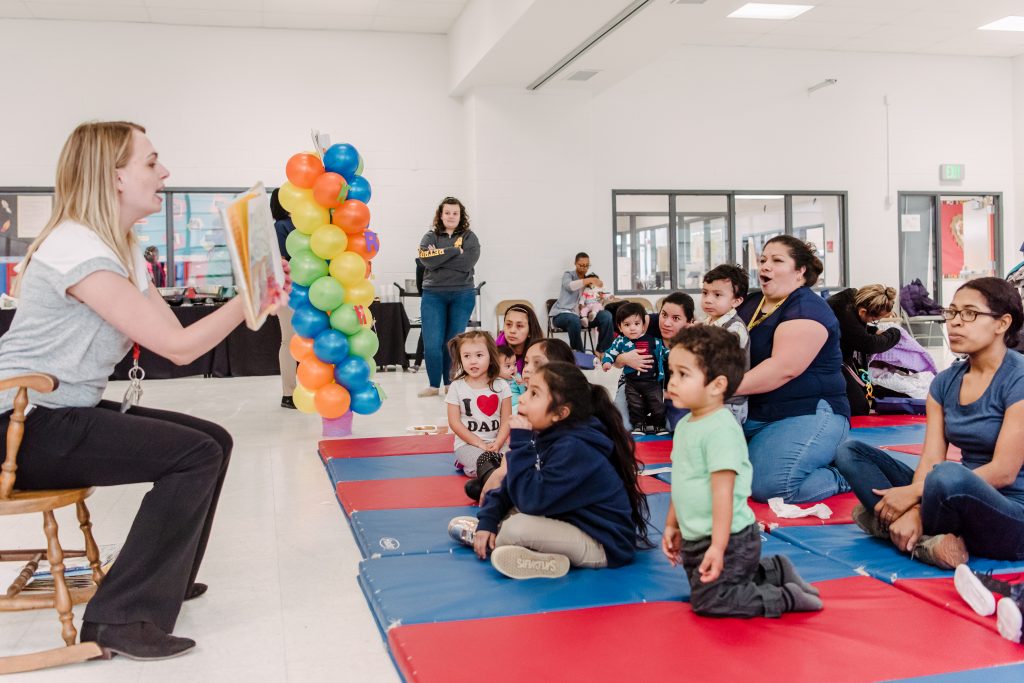 A woman sits and holds up a children's book and faces a group of young children and their parents who are sitting on a red and blue mat.