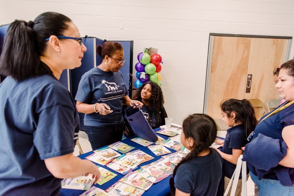 A group of women of different ages and races wearing dark blue t-shirts stand around a table with children's books while two young girls wearing blue shirts stand in front of the table.