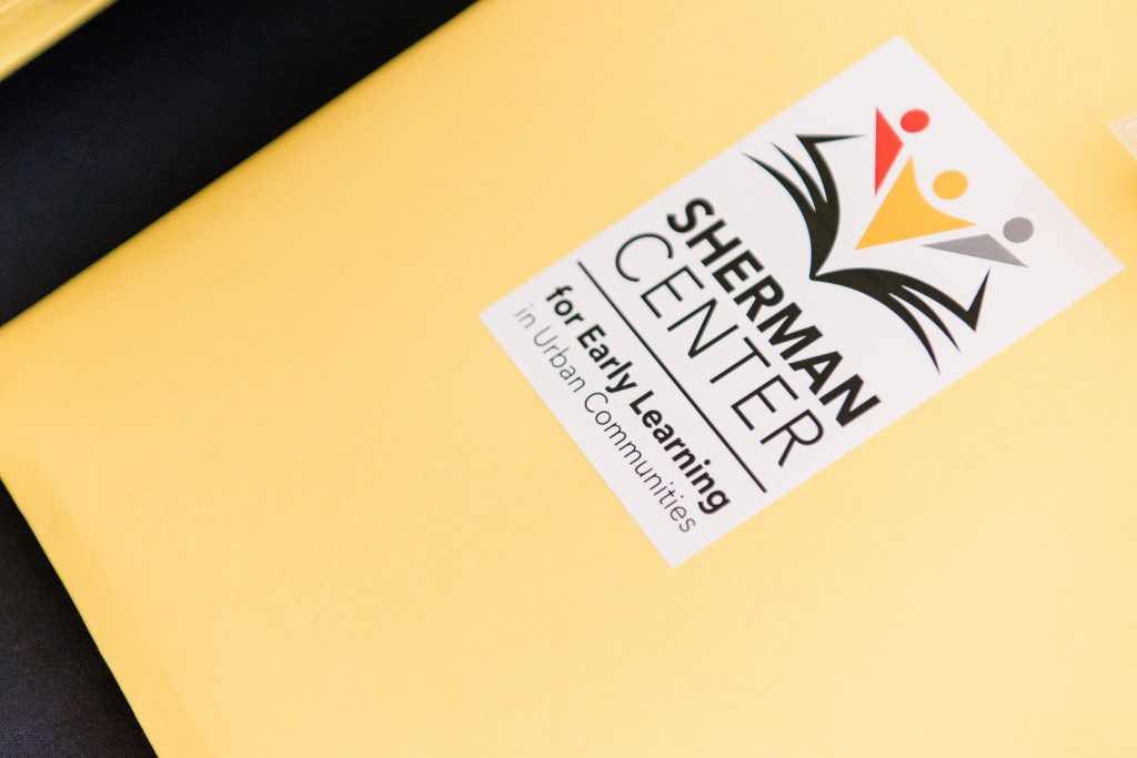 Bright gold paper with a white rectangle sticker in the middle with a logo and the words Sherman Center for Early Learning in Urban Communities written in black.