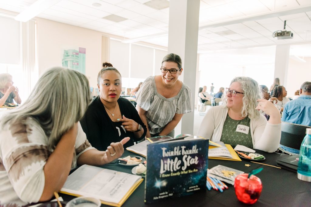 Four women of mixed races and ages have a discussion while seated around a table with books in front of them.