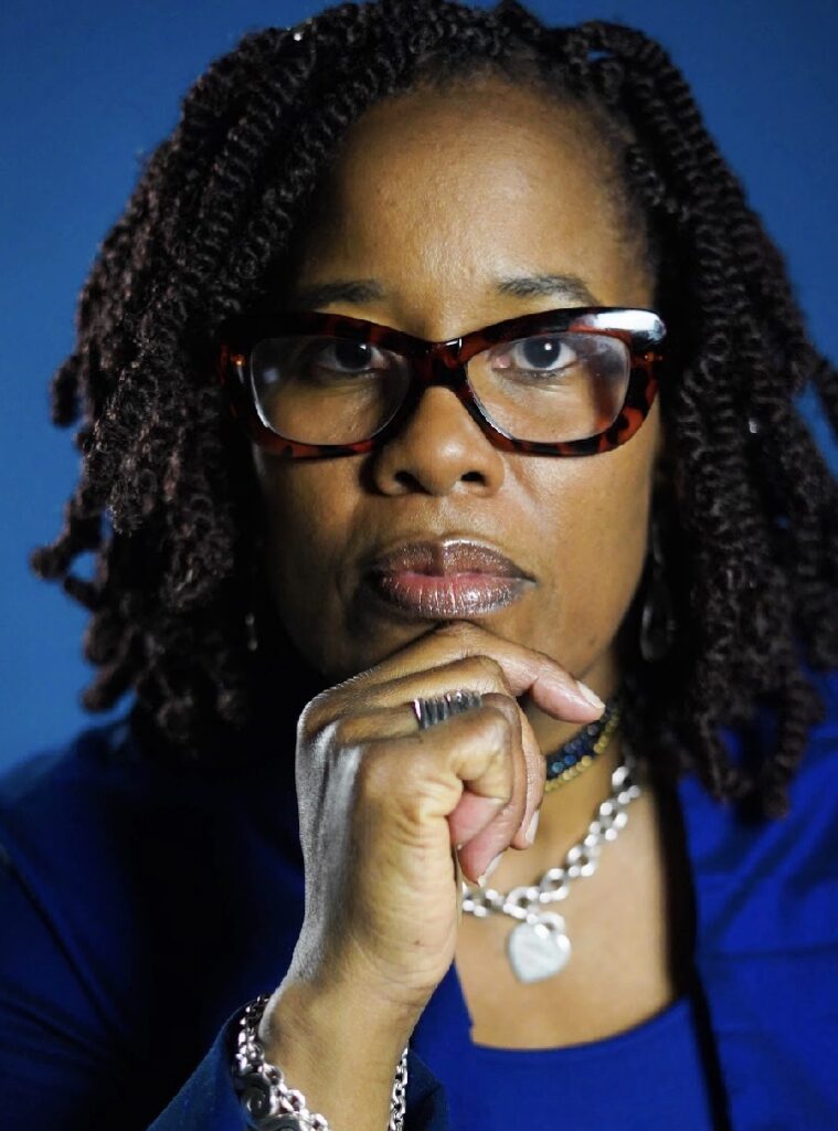 A Black woman with curly black hair wearing dark rimmed glasses and a cobalt blue suit looks straight at the camera holding her hand up to her chin.